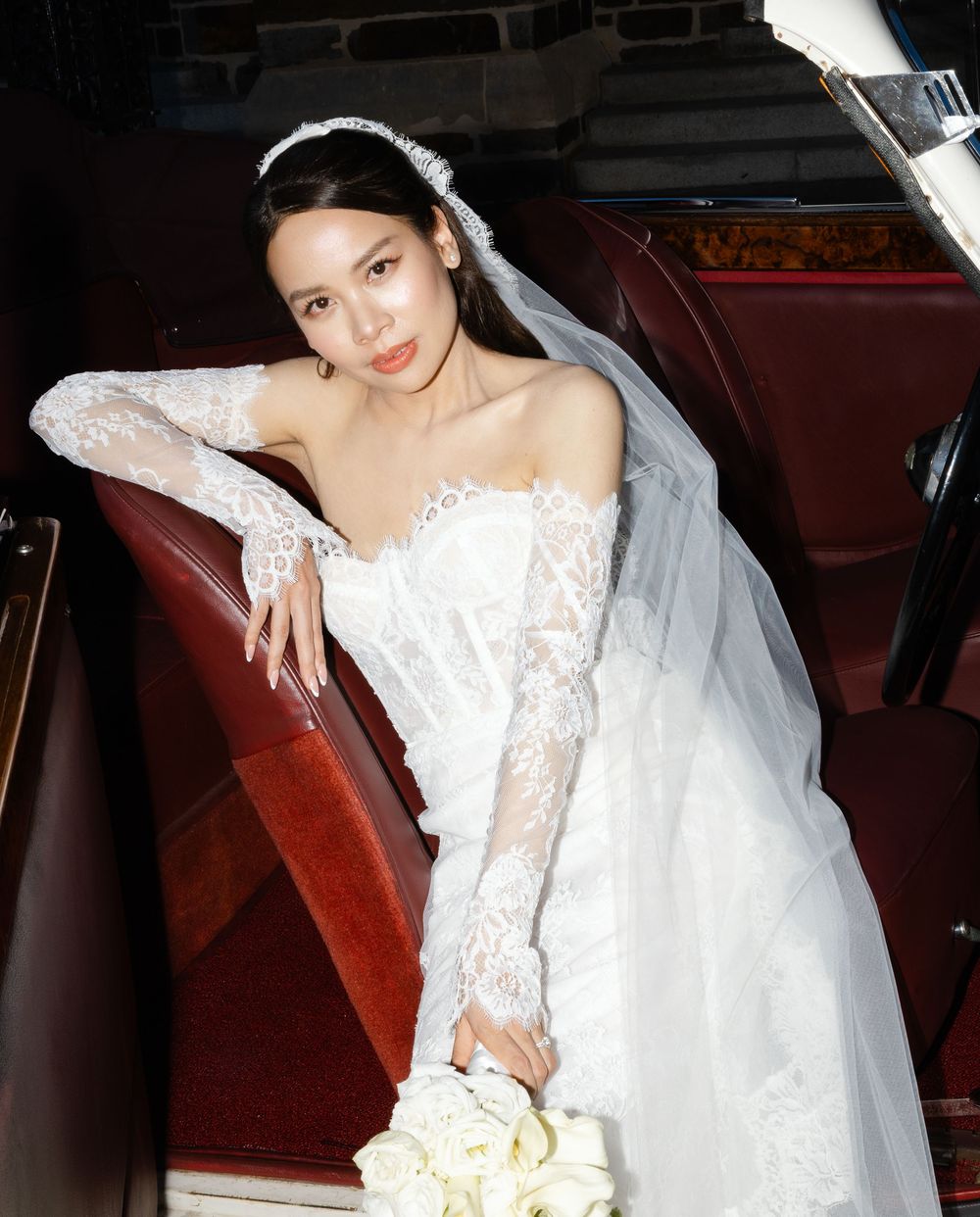 A bride in an elegant white lace gown sits gracefully in a vintage car interior.