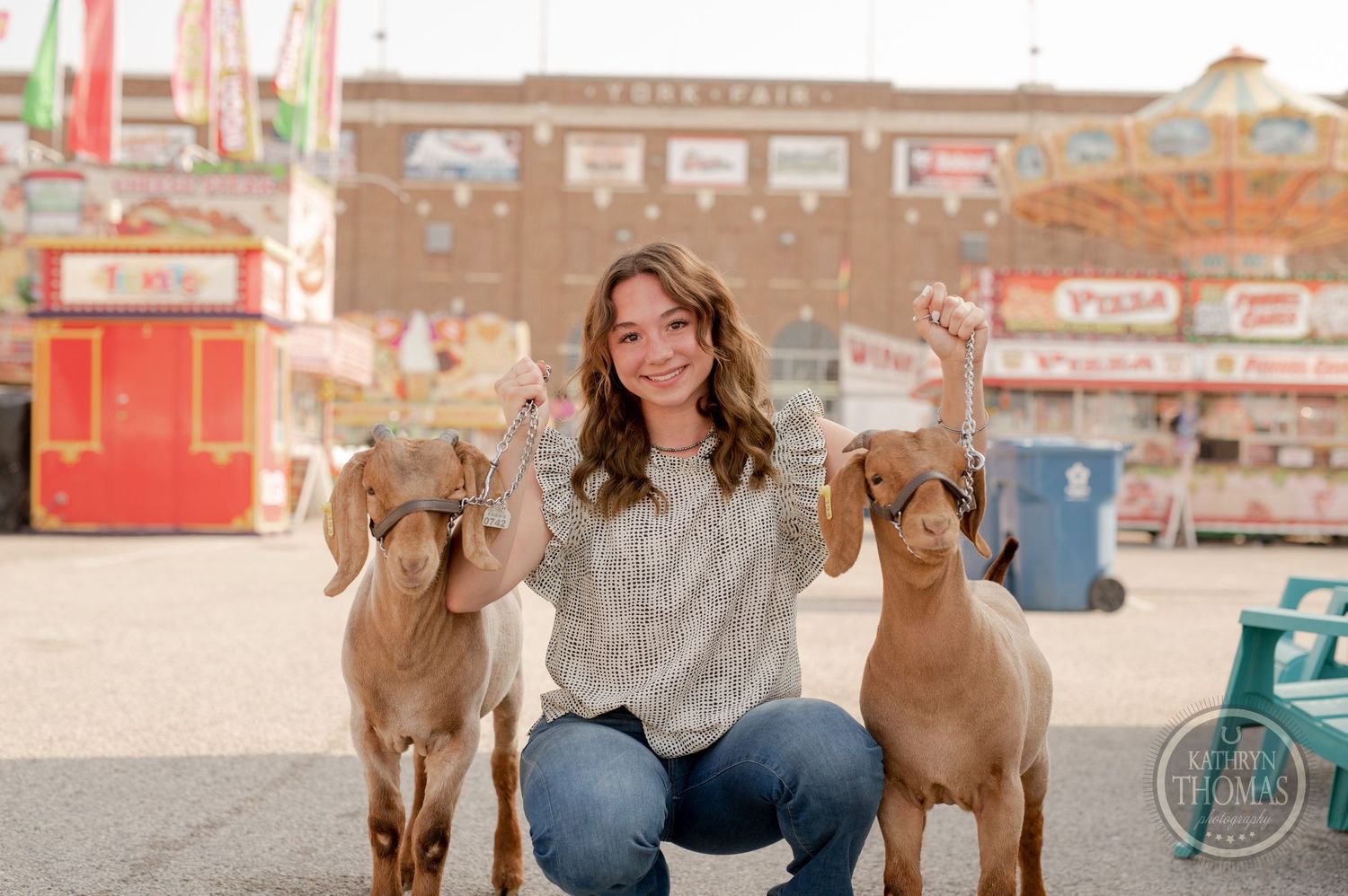 Two tan goats pose with handler at outdoor carnival setting with food stands in background.
