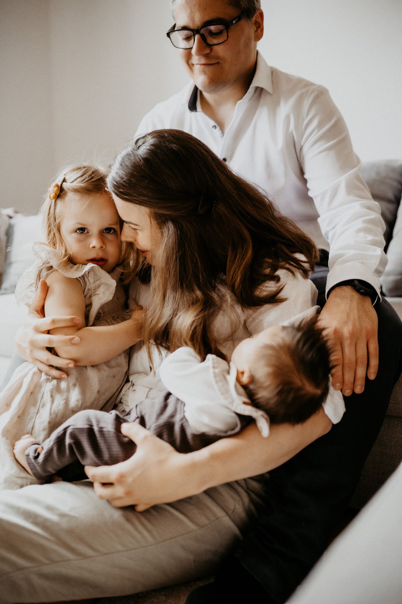 Intimate family moment on grey couch showing parents cuddling with their baby and toddler.