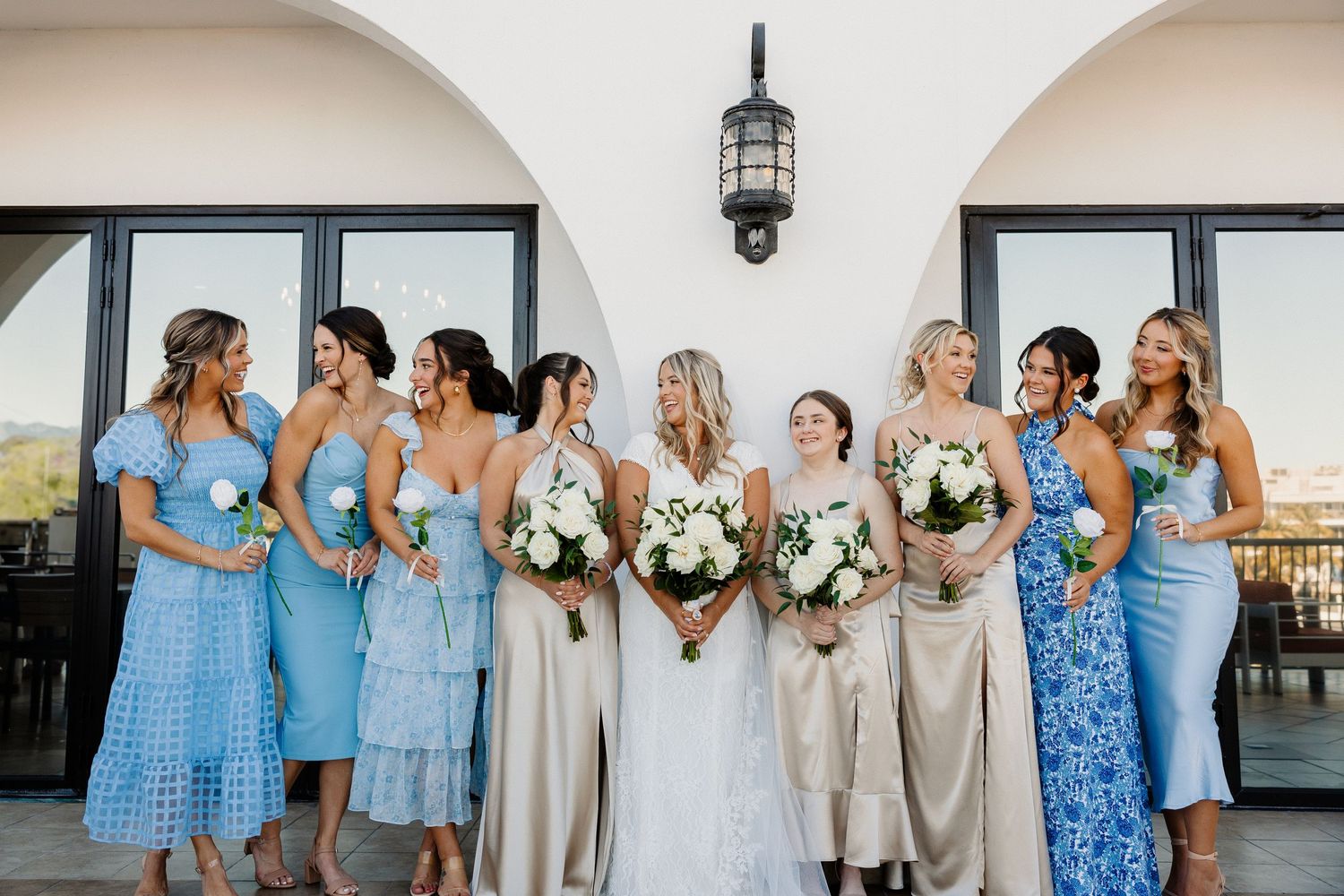 Bride poses with bridesmaids in blue and champagne dresses holding white bouquets outdoors.