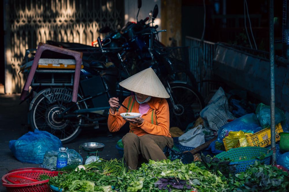 Street vendor in conical hat selling fresh vegetables at a local market stall.