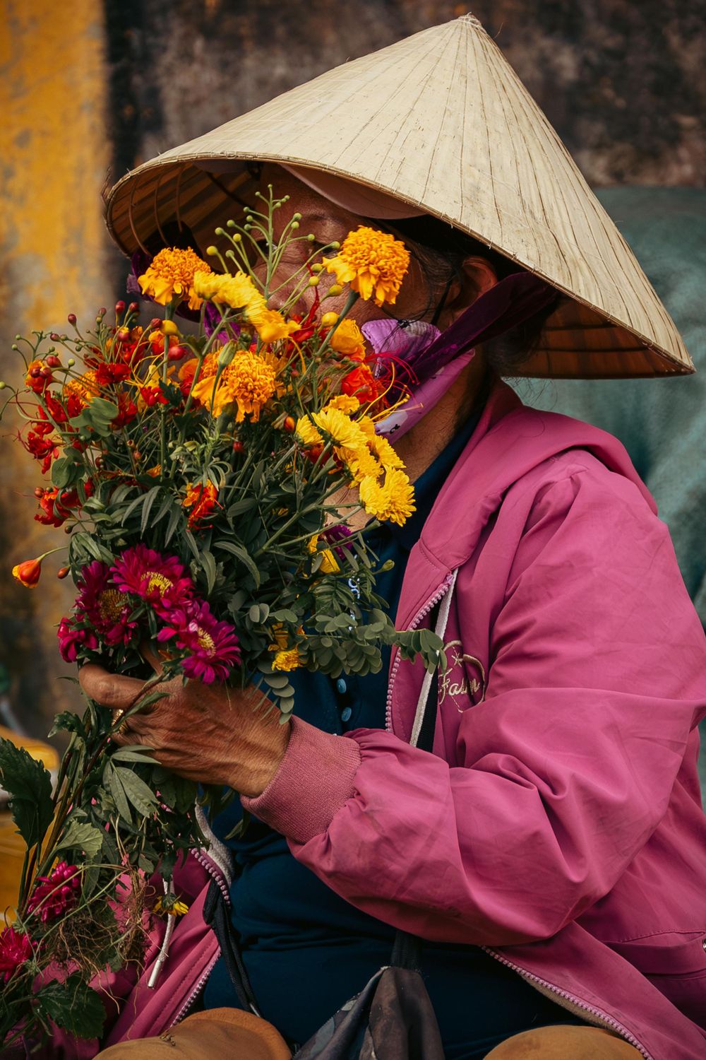 Flower vendor wearing conical hat and pink jacket holds colorful bouquet of marigolds and chrysanthemums.