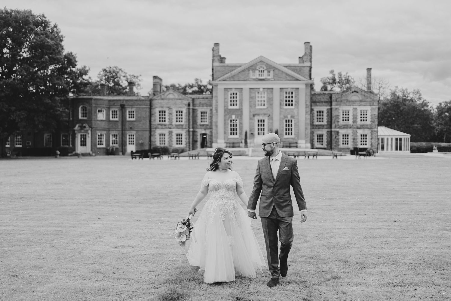 A black and white wedding photo captures a couple walking away from a grand manor house.