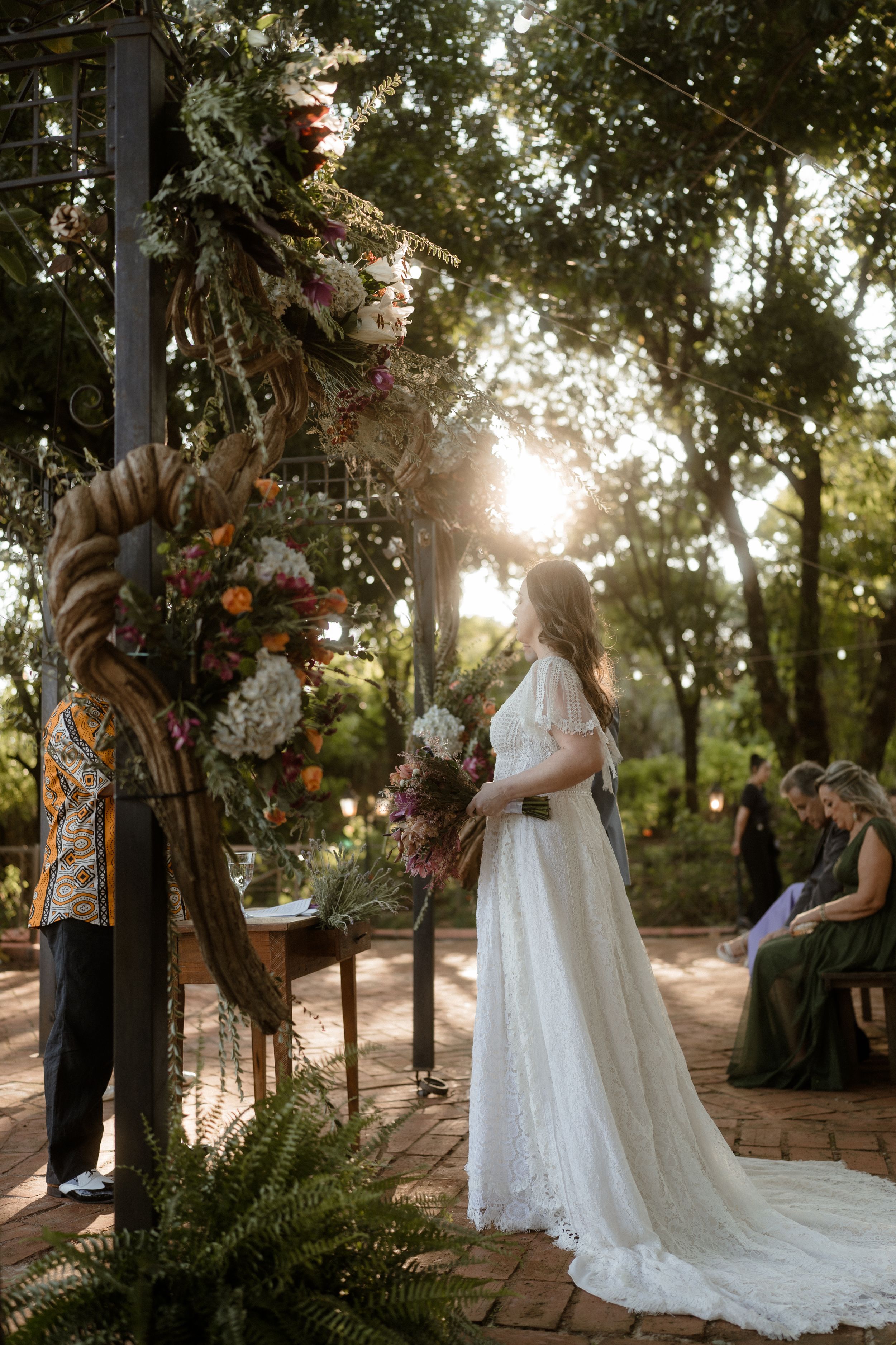 Outdoor wedding ceremony with a wooden arch decorated in autumn florals and foliage as sunlight streams through trees.