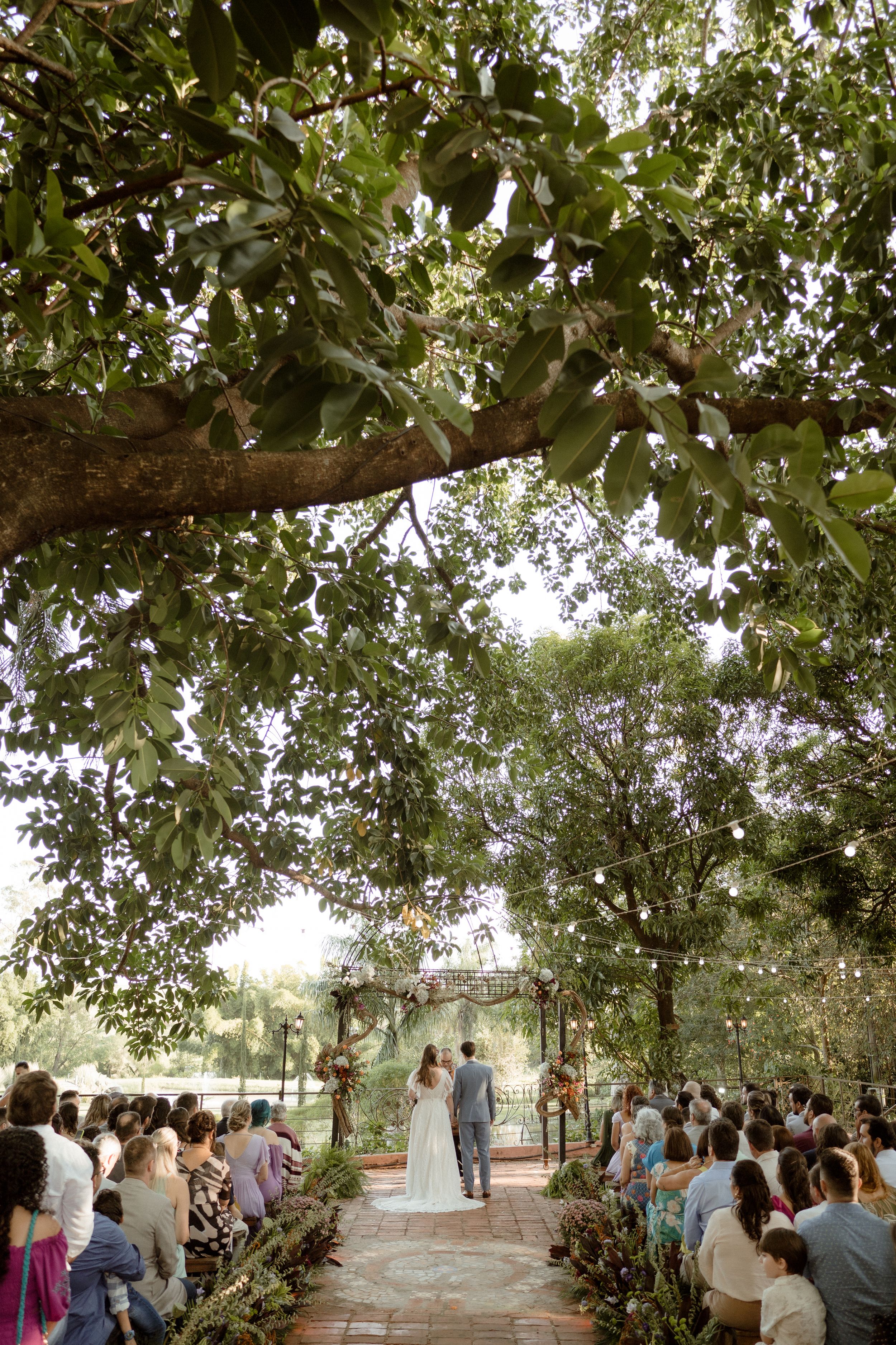 Wide shot of wedding ceremony under tree canopy with guests seated along stone pathway.