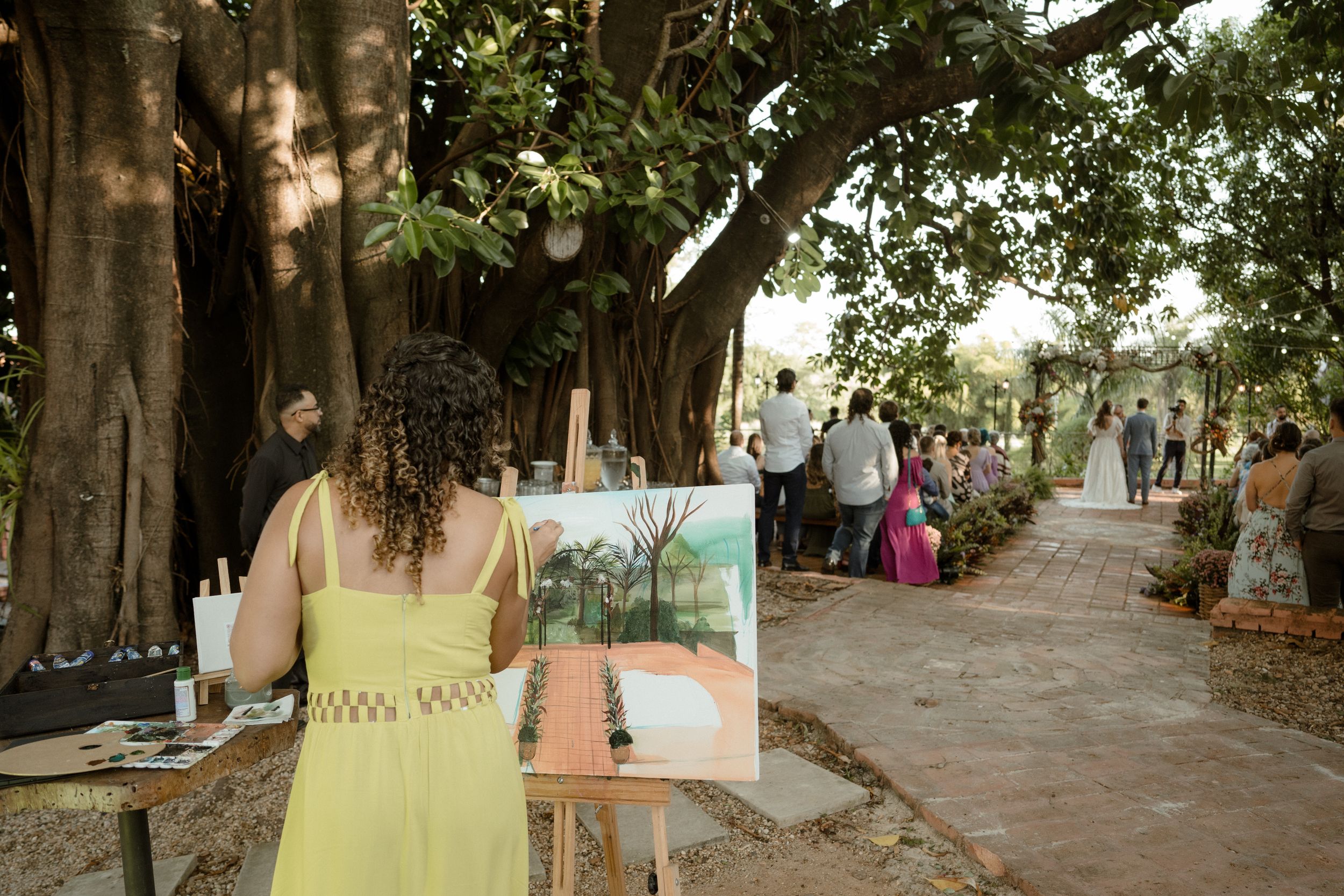 Artist painting a watercolor scene during outdoor event with trees and pathway in background.