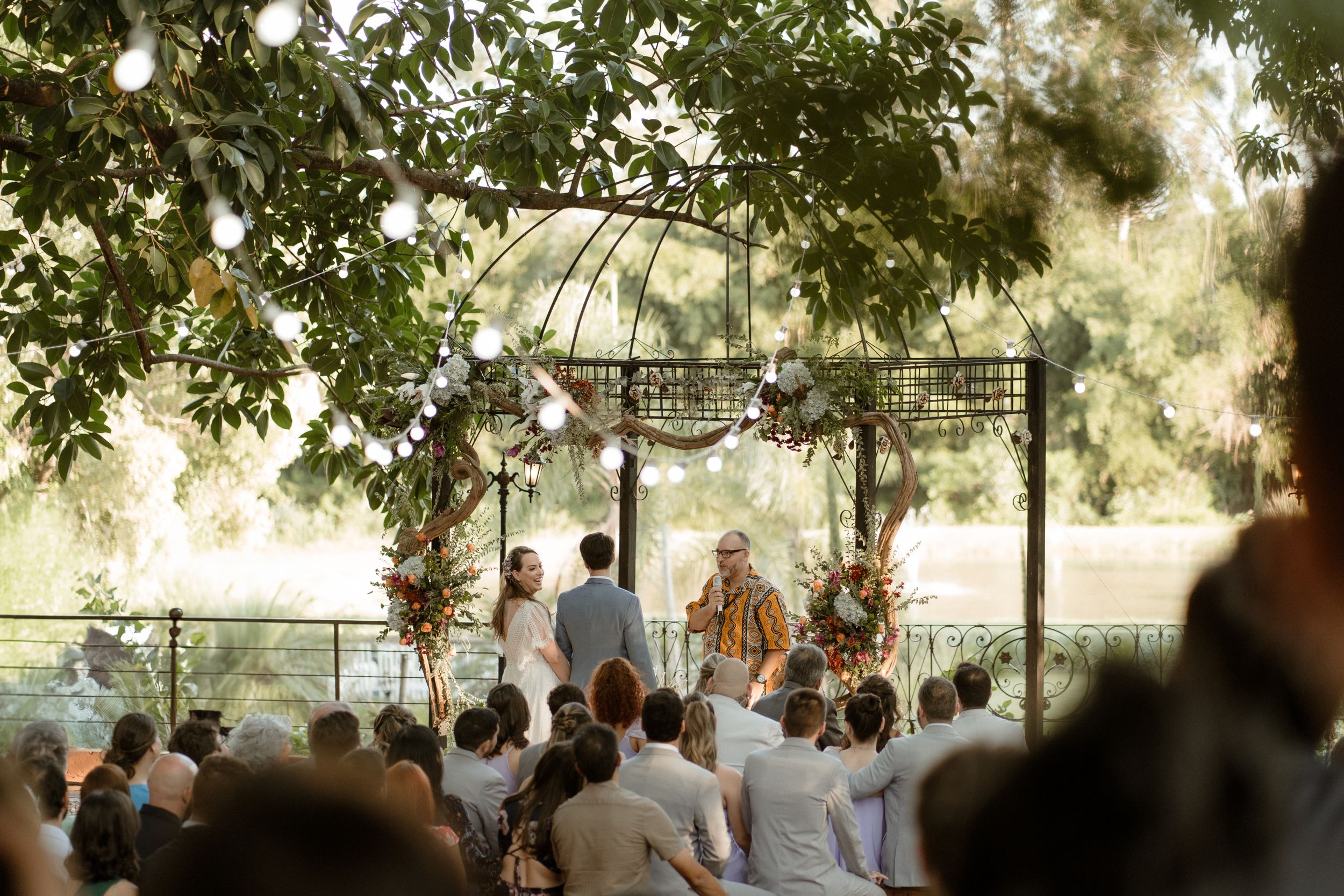 Outdoor wedding ceremony under metal gazebo with natural tree canopy and string lights.
