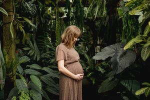 An expectant woman in a brown dress stands serenely among lush tropical plants in a greenhouse setting.