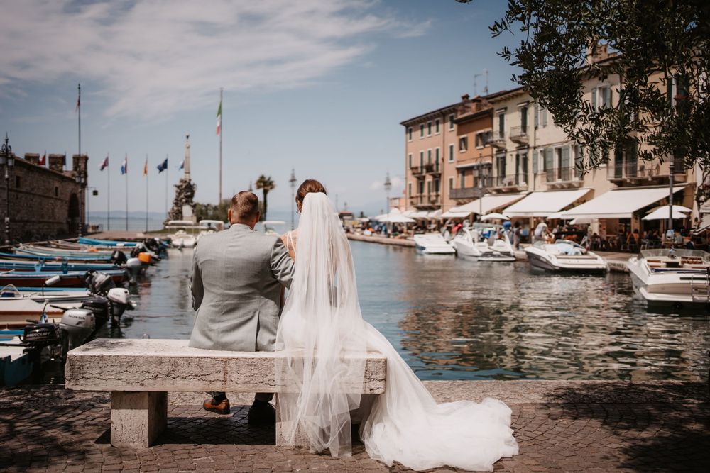 Couple sits on bench overlooking Mediterranean marina with boats and buildings in romantic wedding setting.