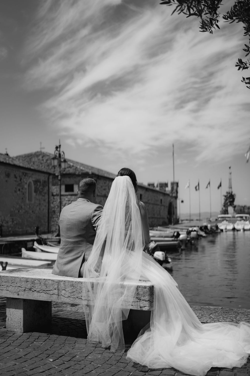 Black and white wedding photo of couple sitting by harbor with flowing veil.