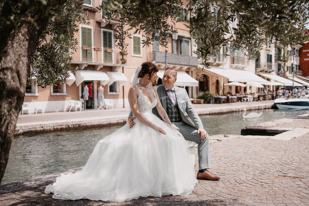Wedding couple seated on waterfront promenade with classic European buildings and boats in background.