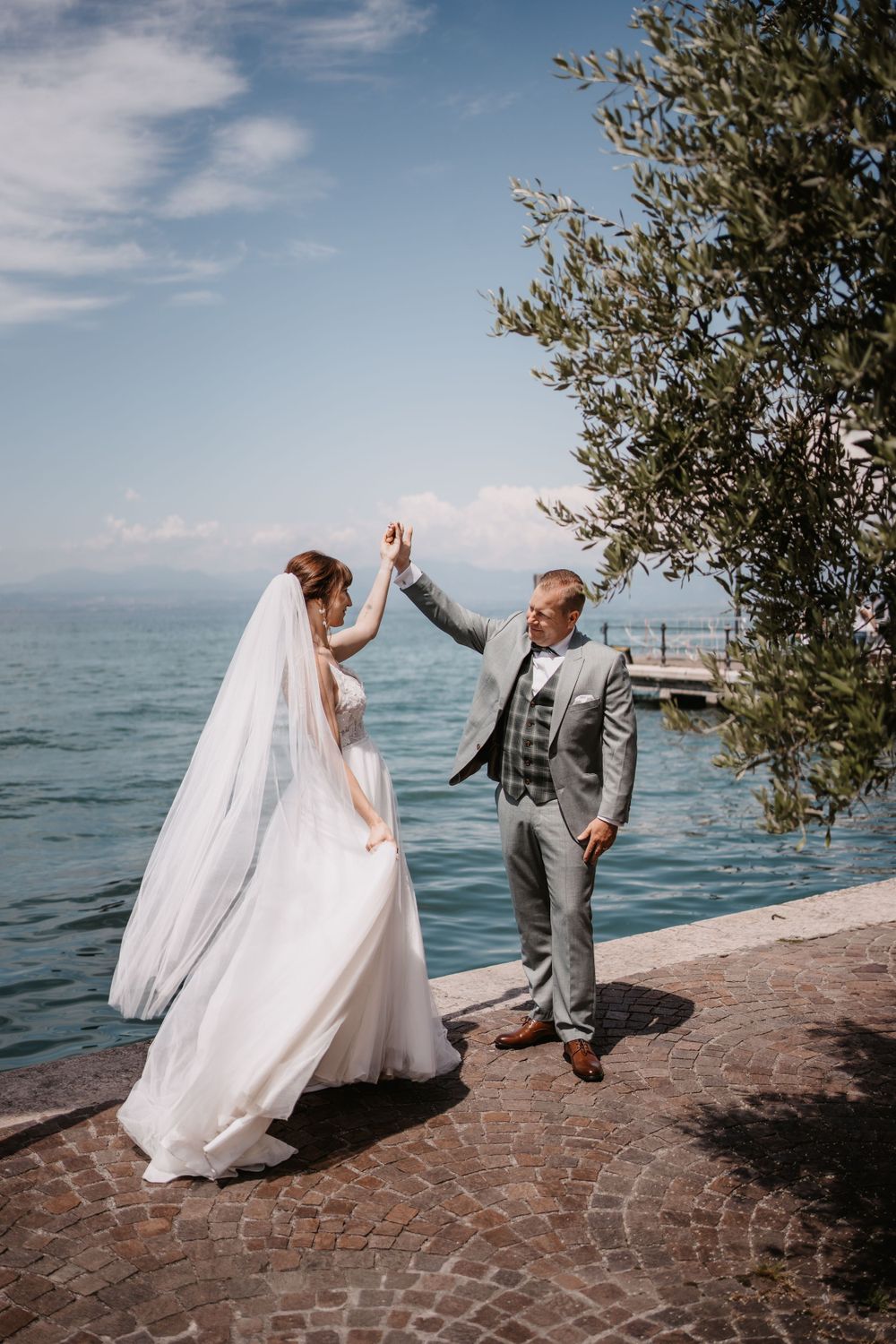 Wedding couple sharing celebratory high-five by Mediterranean waterfront with olive tree.