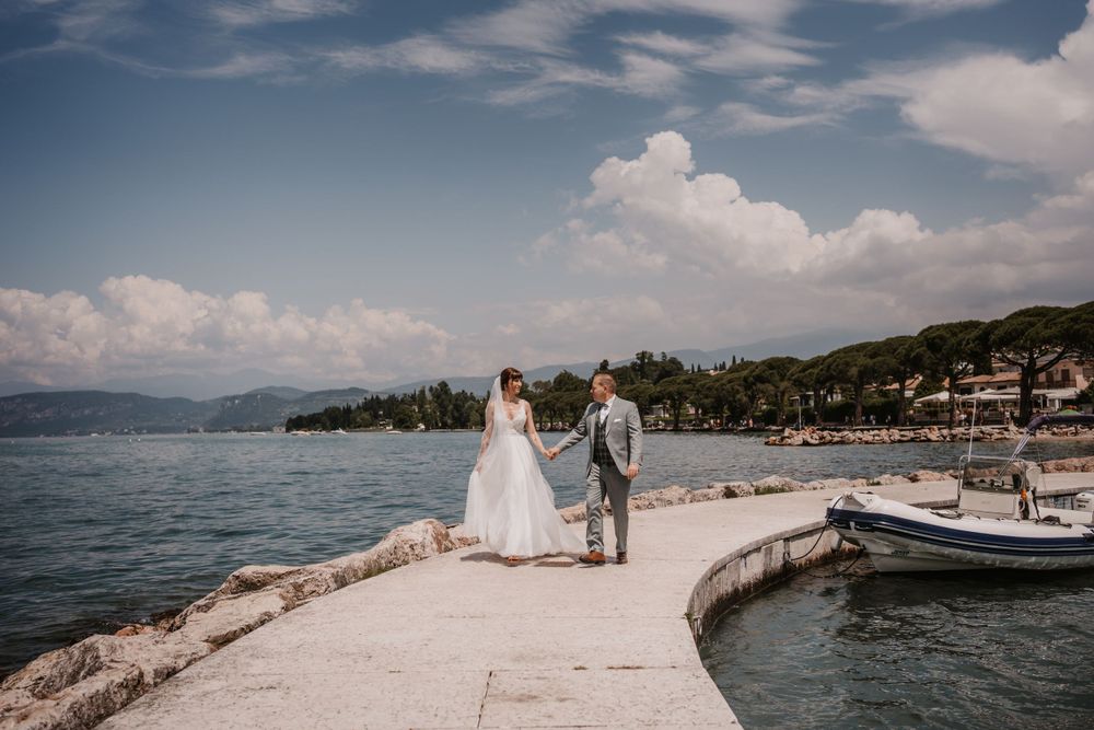 Wedding couple stands on curved marina walkway with boats and coastal scenery.