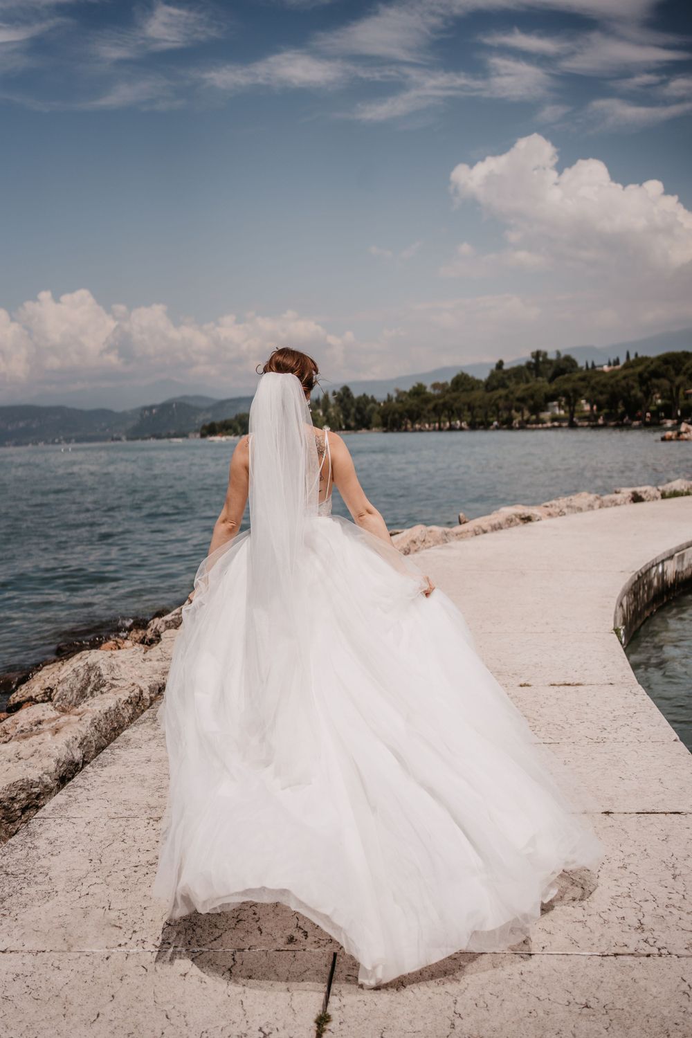Flowing white wedding dress and veil catch breeze on Mediterranean waterfront.