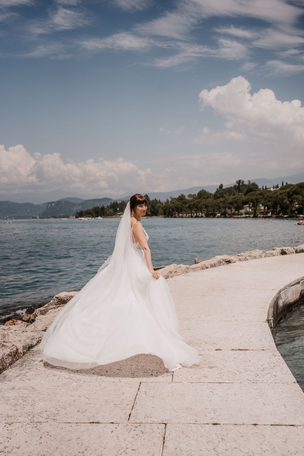 Bride in flowing gown poses on coastal promenade with mountain and sea views.