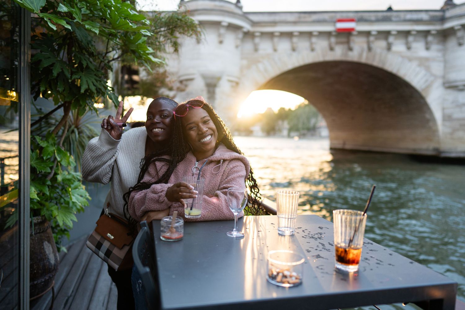 Two people enjoying drinks at a riverside cafe table in Paris with a stone bridge and sunset in the background.