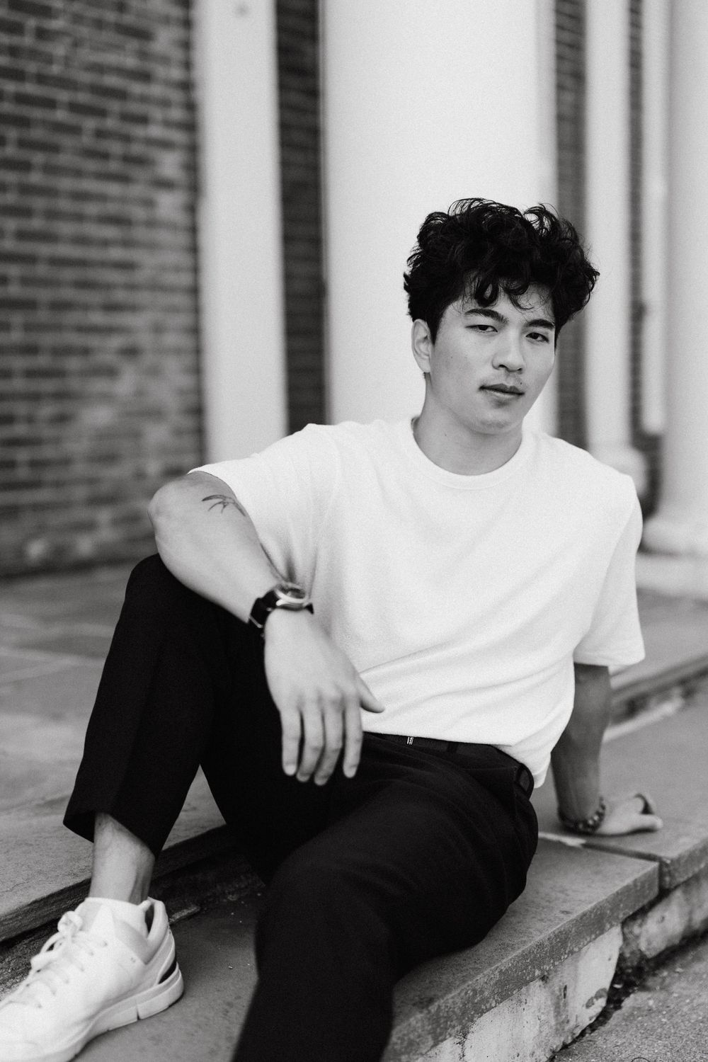 Black and white portrait of a young man with curly dark hair in a white t-shirt and black pants, sitting on brick steps near white columns.