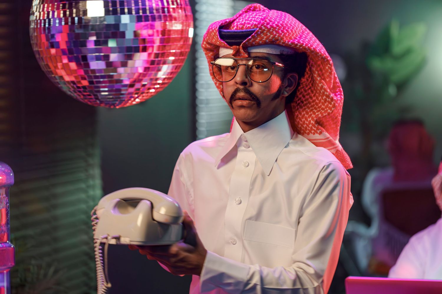 Someone in traditional Middle Eastern dress and glasses stands beneath a disco ball in a nightclub setting.