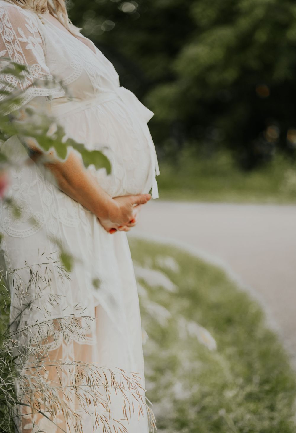 Side view of a pregnant woman in a white lace dress standing in a garden setting.
