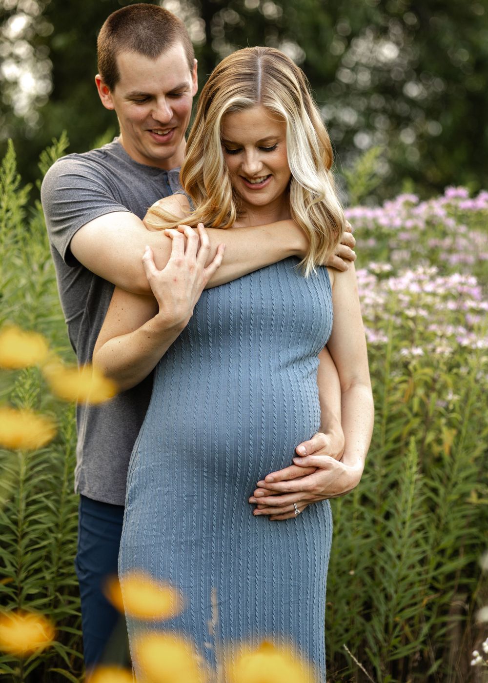 Couple embracing in a flower garden with woman wearing a blue knit maternity dress.
