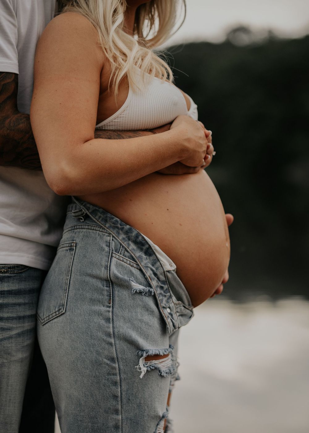 Side view of pregnant belly in ripped jeans with partner's hands showing tender embrace.