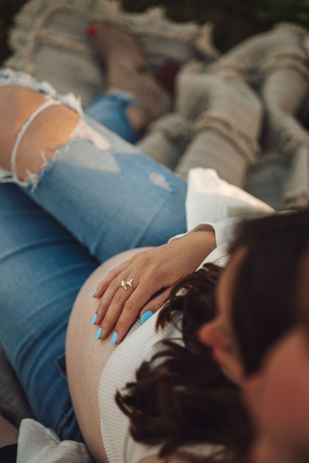 Close-up of a hand with an engagement ring resting on a pregnant belly in ripped jeans.