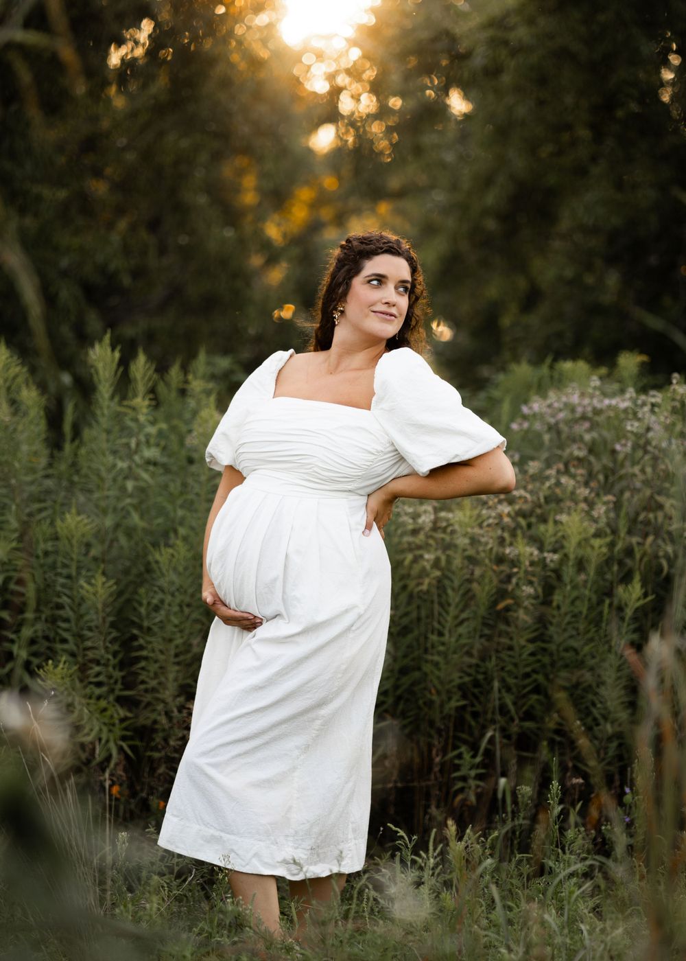 Expectant mother in a white dress poses in a garden during golden hour sunset.