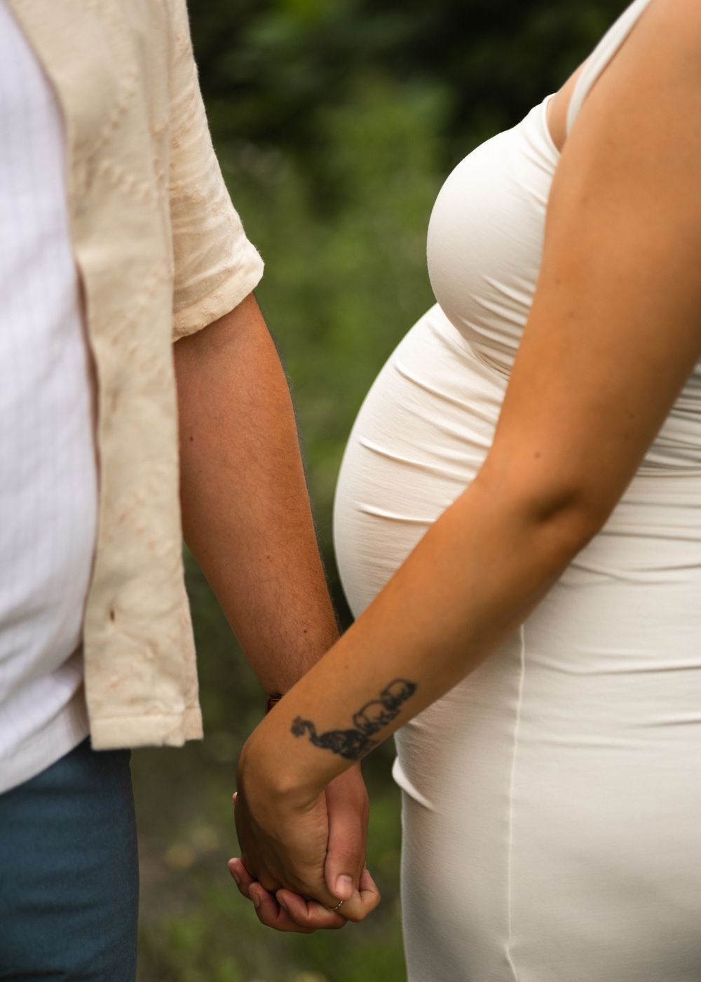 Close-up of couple holding hands, showing a small tattoo and white maternity dress.