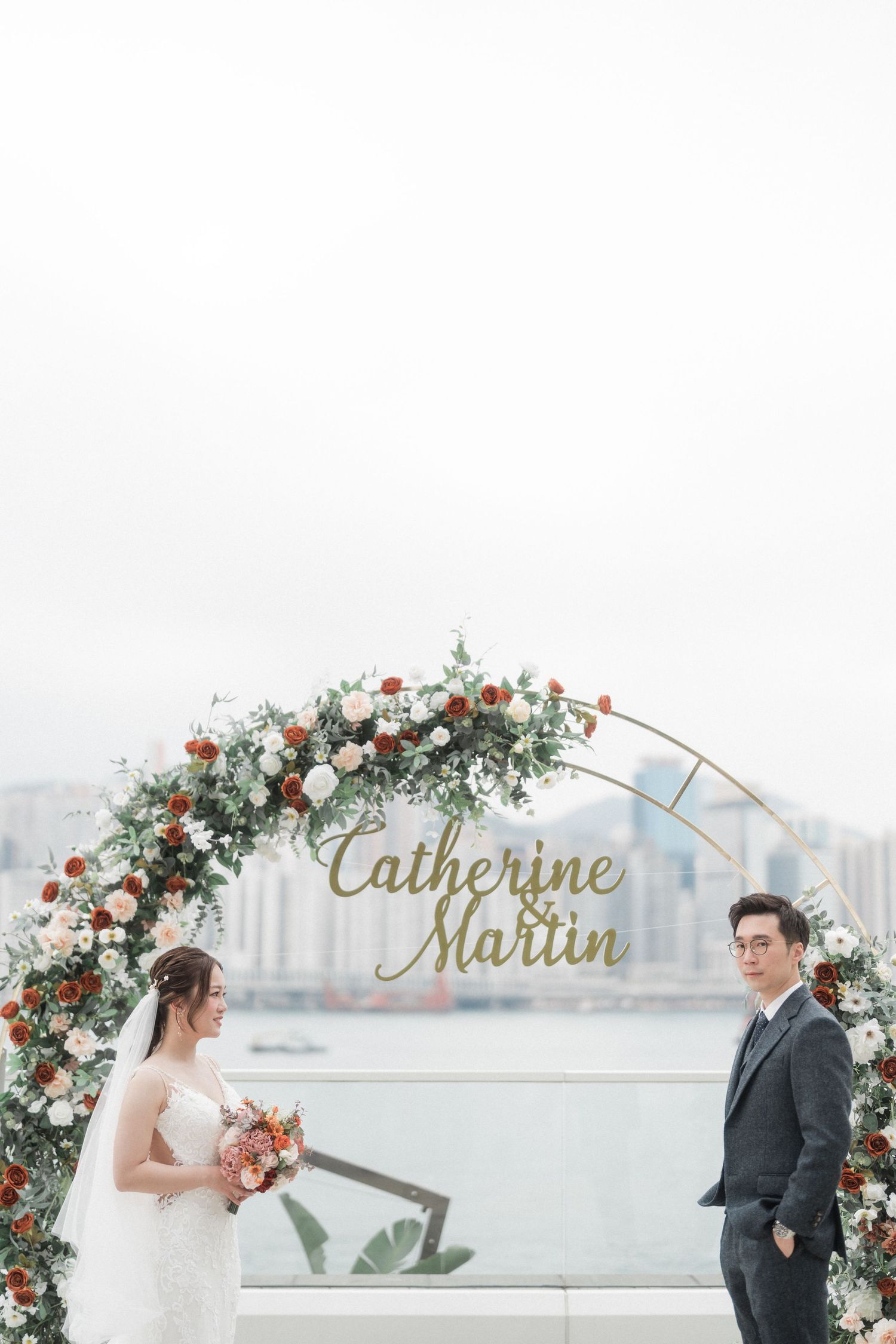Wedding arch decorated with pink and white flowers frames a couple against a cityscape.