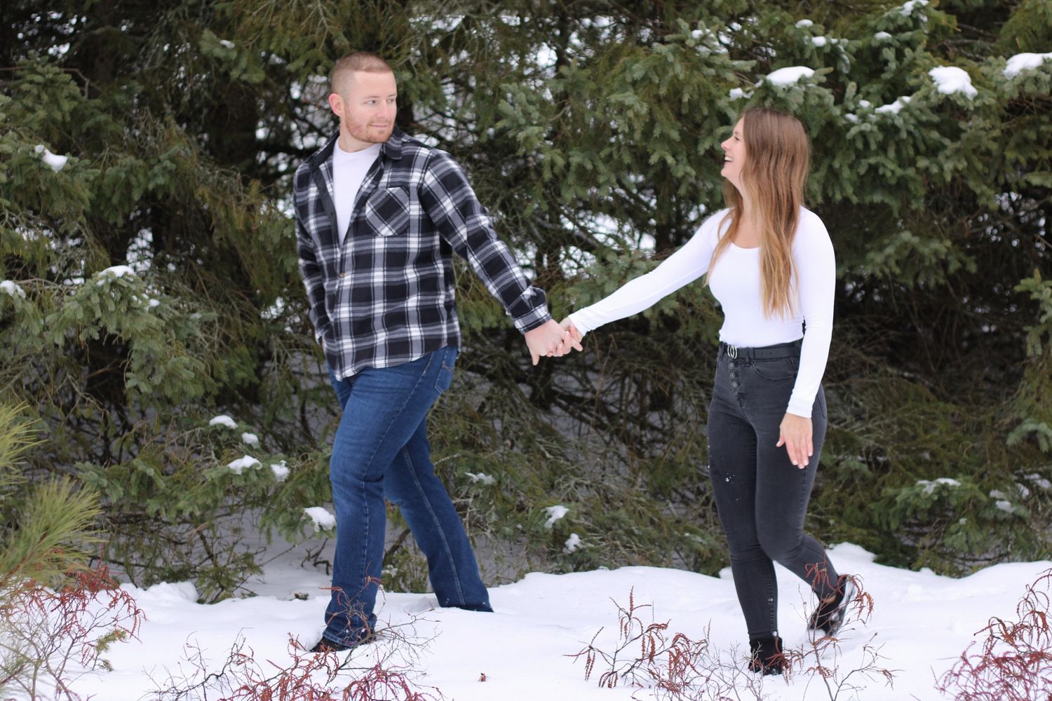 Couple holding hands walking through a snowy winter landscape with evergreen trees in the background.