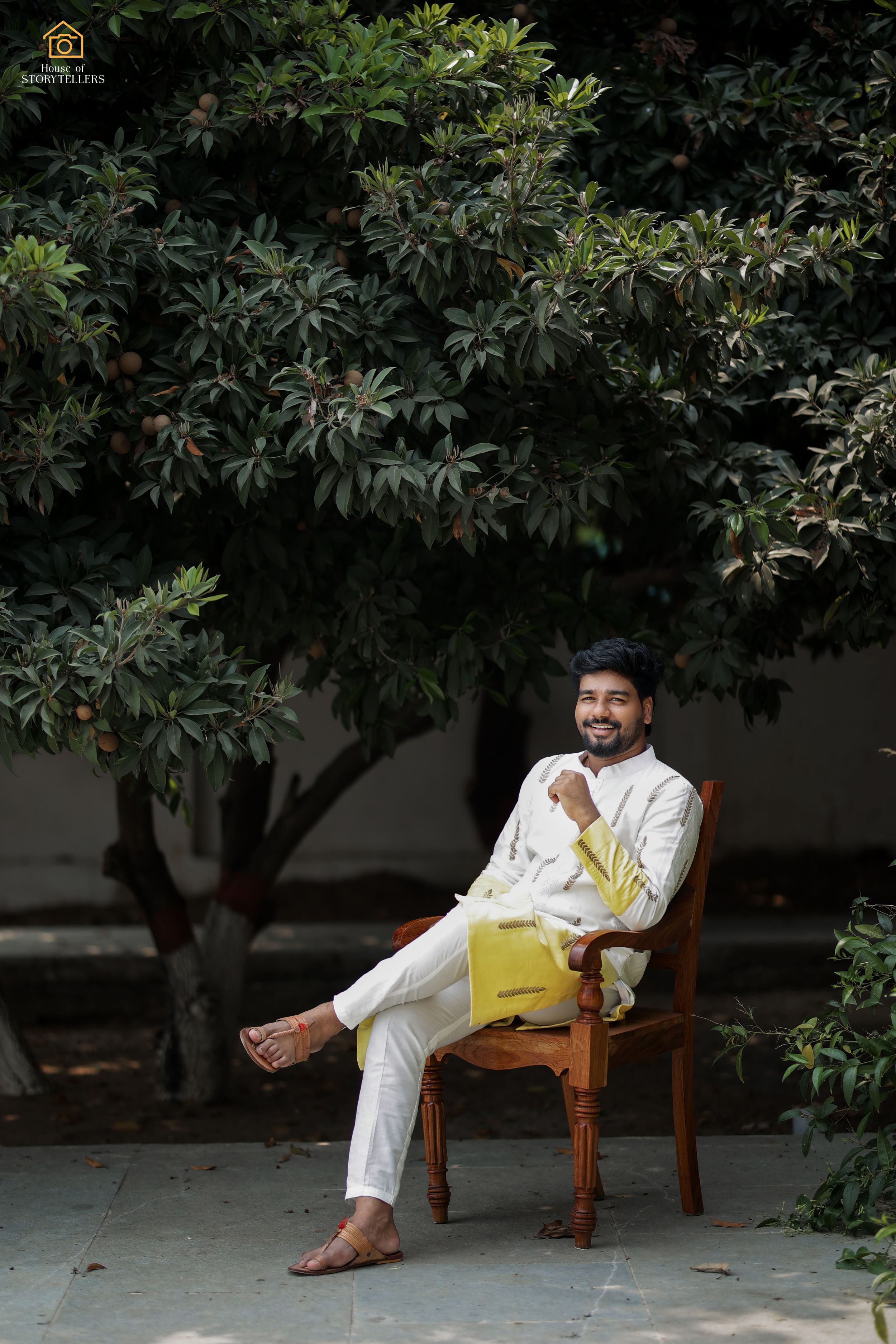 Person in white kurta sitting casually on wooden chair under lush tree.