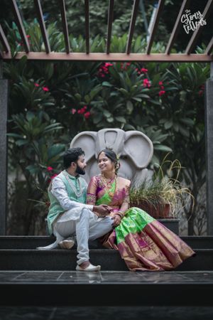 Couple in traditional attire sitting on bench surrounded by tropical plants and flowers.