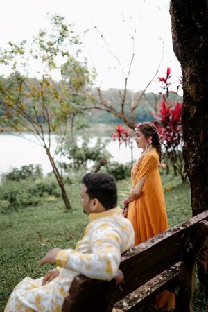 Indian couple in traditional attire near a lakeside garden, bride in orange dress and groom in white outfit on a bench.