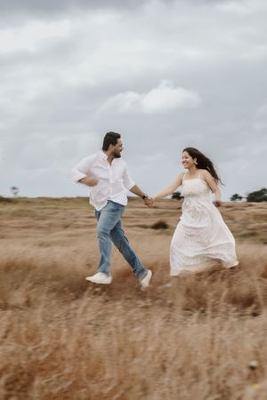 A joyful couple holds hands while running through a golden field, smiling at each other on a cloudy day.