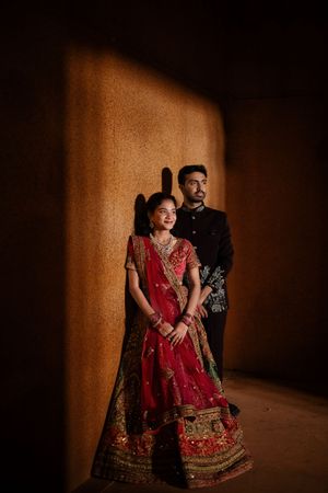 Indian bride in red lehenga and groom in black sherwani pose elegantly against a warmly lit wall.