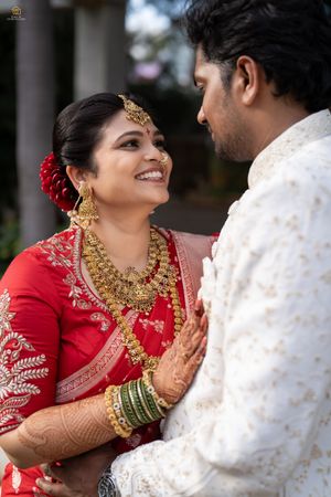 Indian bride in red saree with gold jewelry smiles at groom in white sherwani during traditional wedding ceremony.