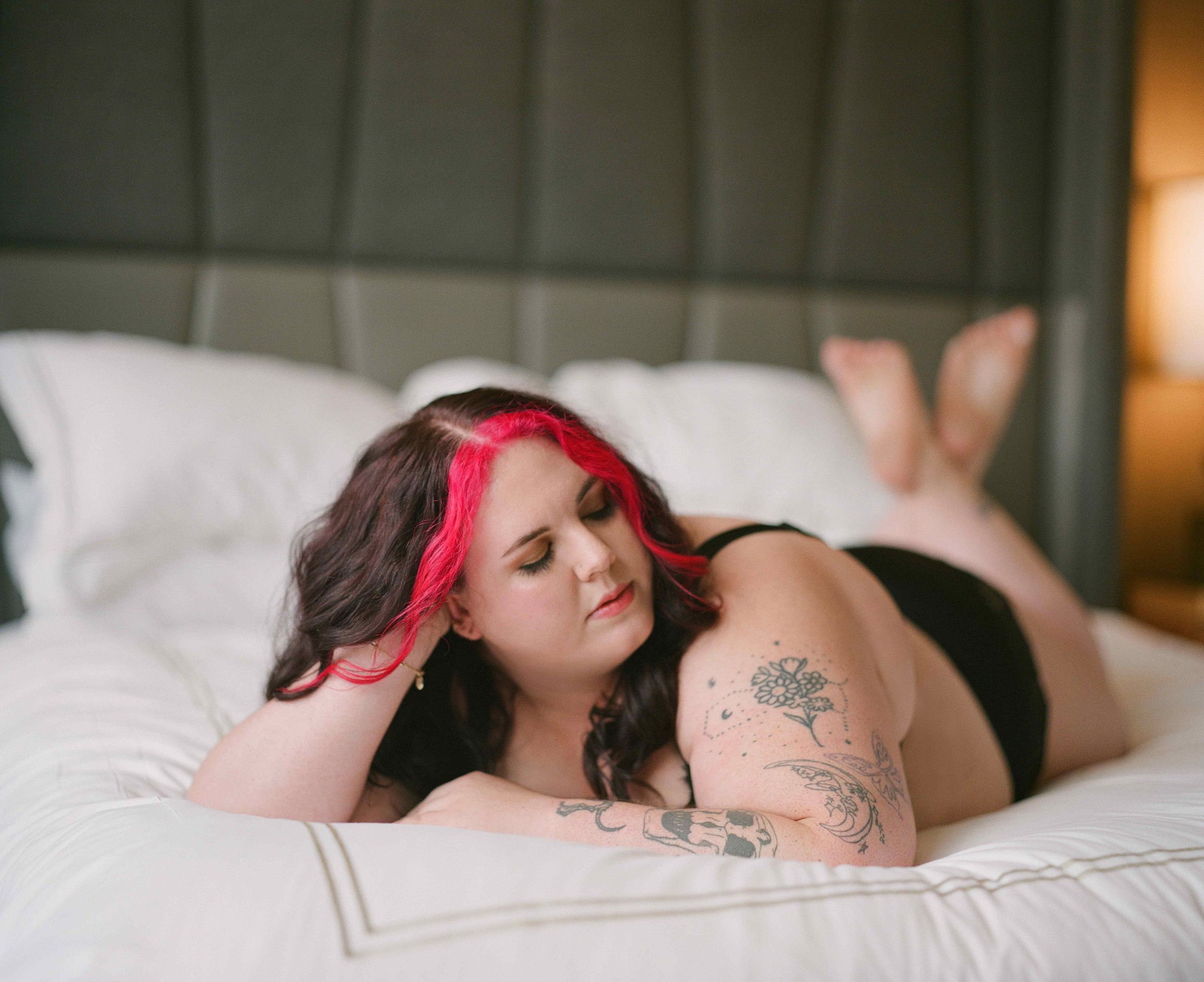 Curvy woman with tattoos wearing red headband relaxes on white bedding in intimate portrait.