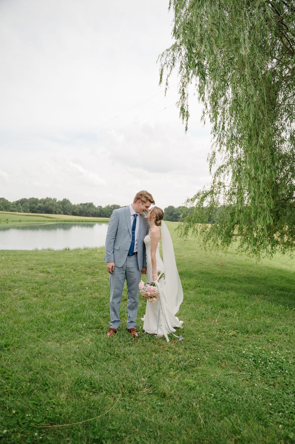 Couple kissing by weeping willow tree and pond on wedding day