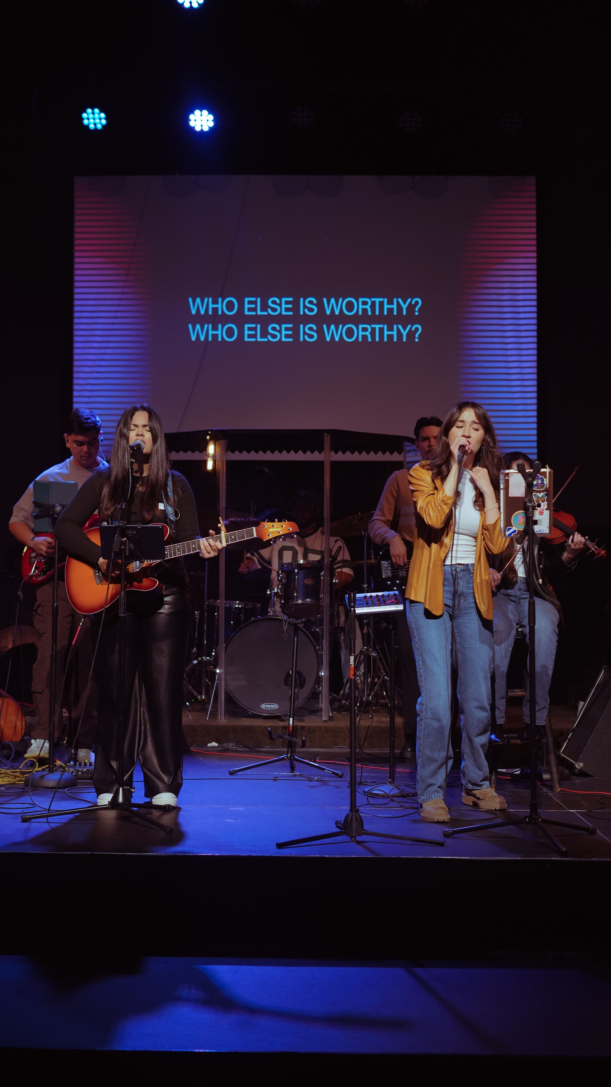 Musicians perform on stage with guitar and microphone under blue lighting with 'WHO ELSE IS WORTHY?' text displayed above.