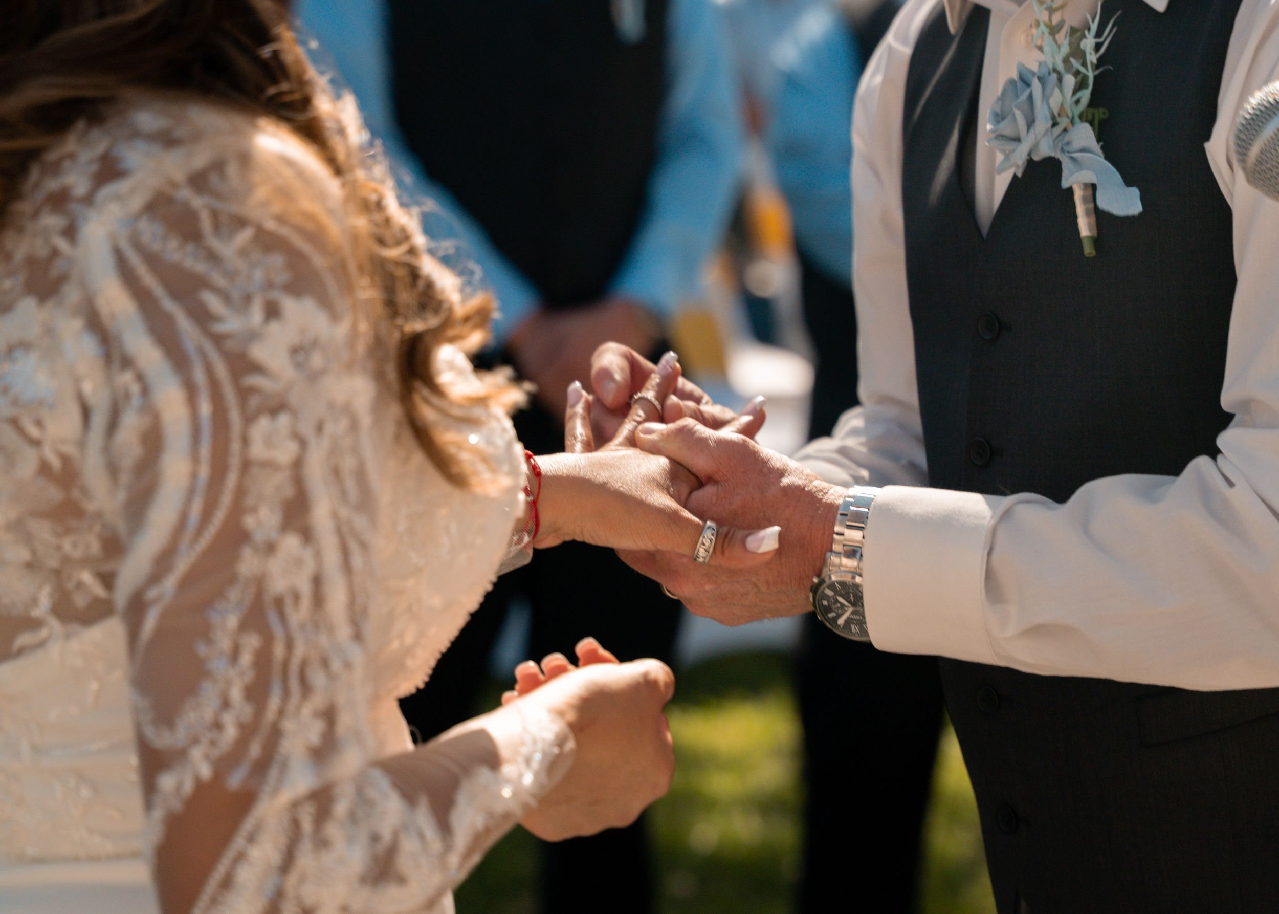 A close-up shot of hands exchanging wedding rings during an outdoor ceremony in bright sunlight.