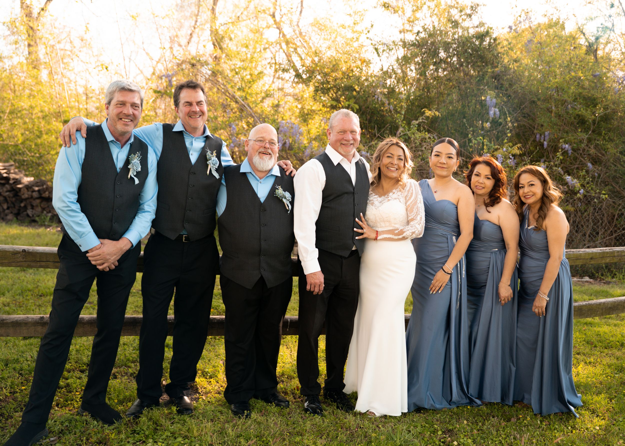 Wedding party in formal attire poses outdoors with men in black vests and bridesmaids in dusty blue dresses at sunset.