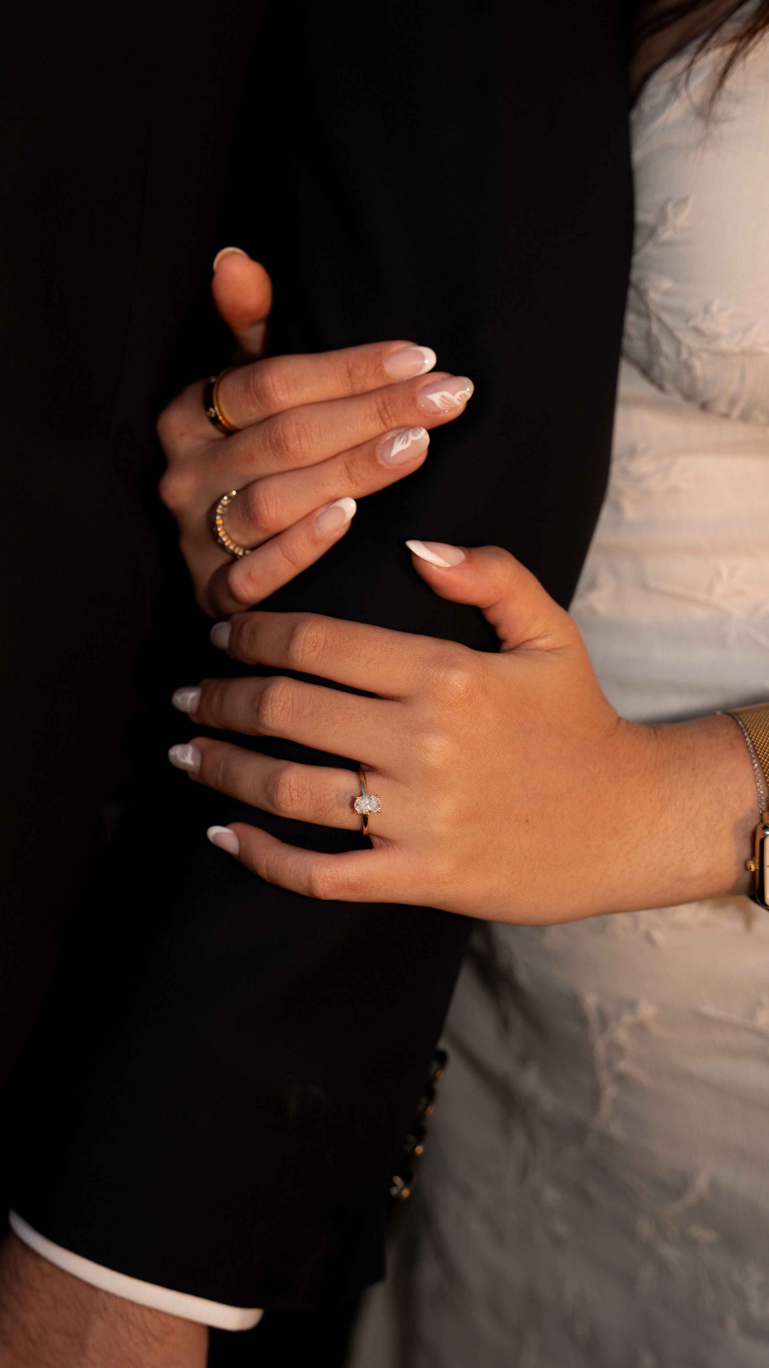 A couple's hands with wedding rings embrace, one wearing a black suit and the other in a white dress.