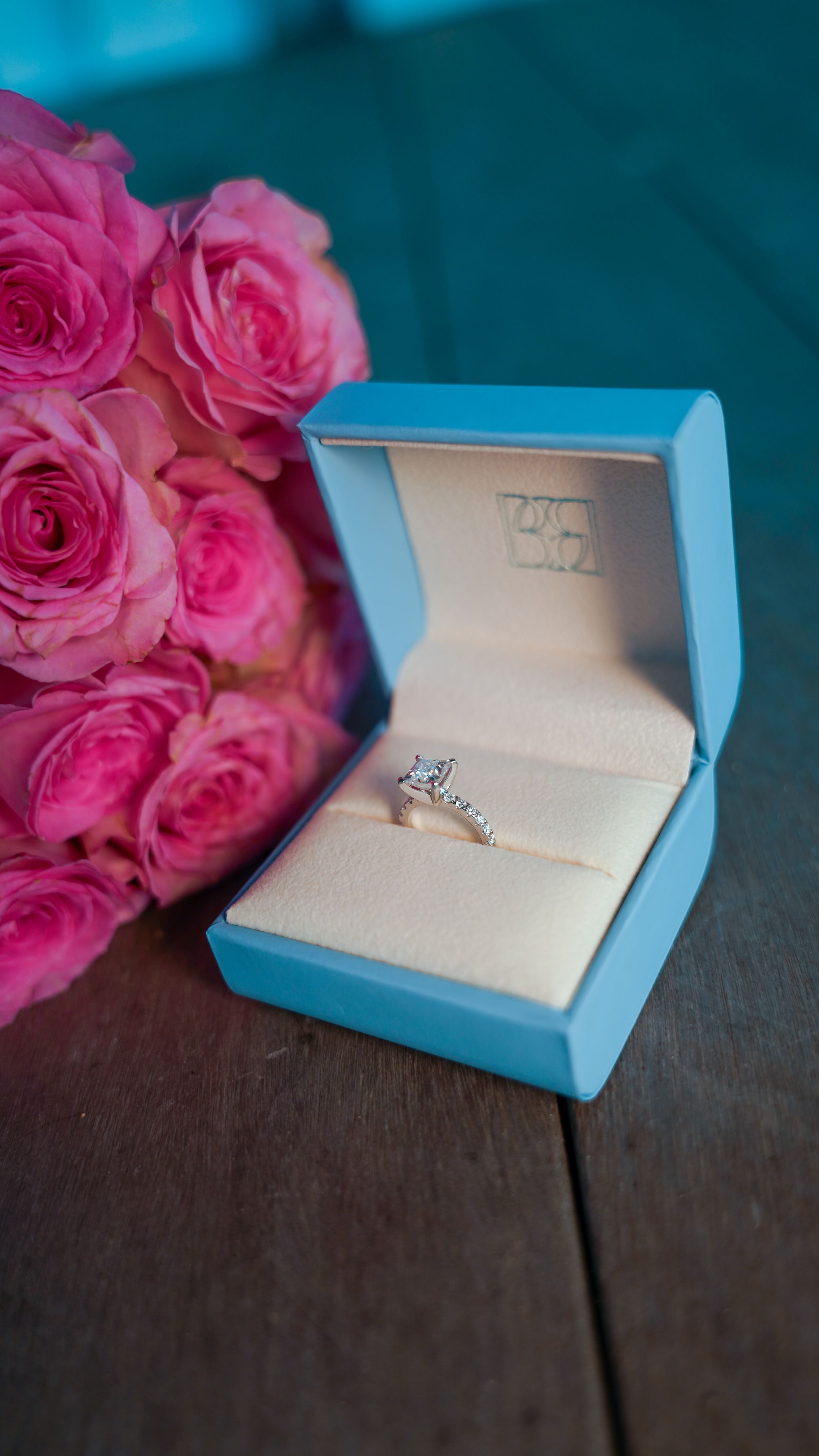 Elegant engagement ring in light blue box with pink roses arranged beautifully on dark wooden surface.