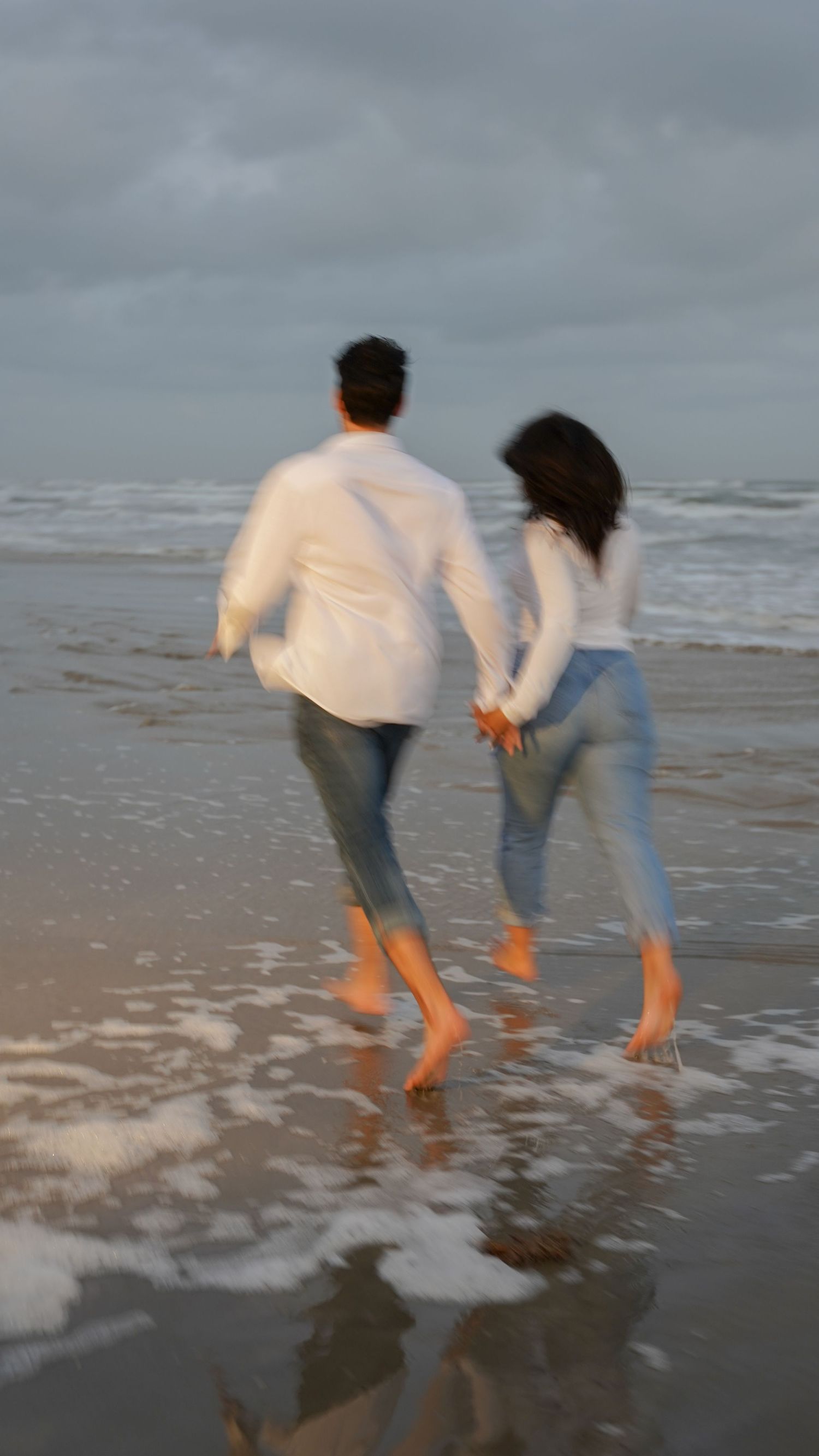 A couple walking barefoot on a beach holding hands at dusk.