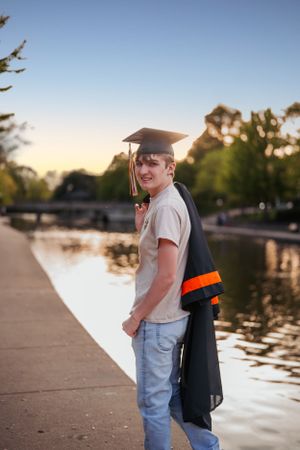 Graduate in cap and gown poses by waterfront at sunset.