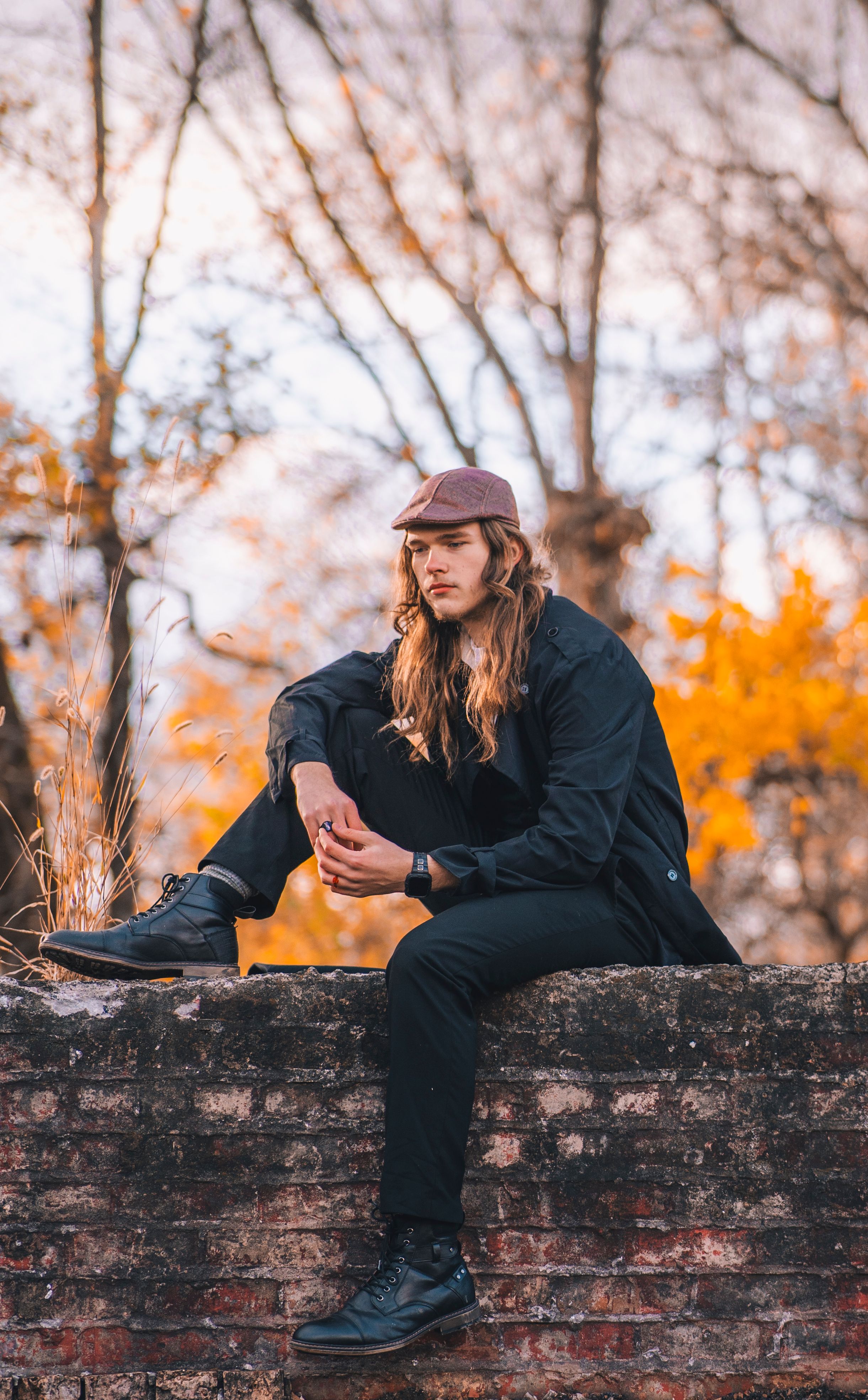 A person in casual attire sits contemplatively on an old brick wall with autumn foliage in the background.