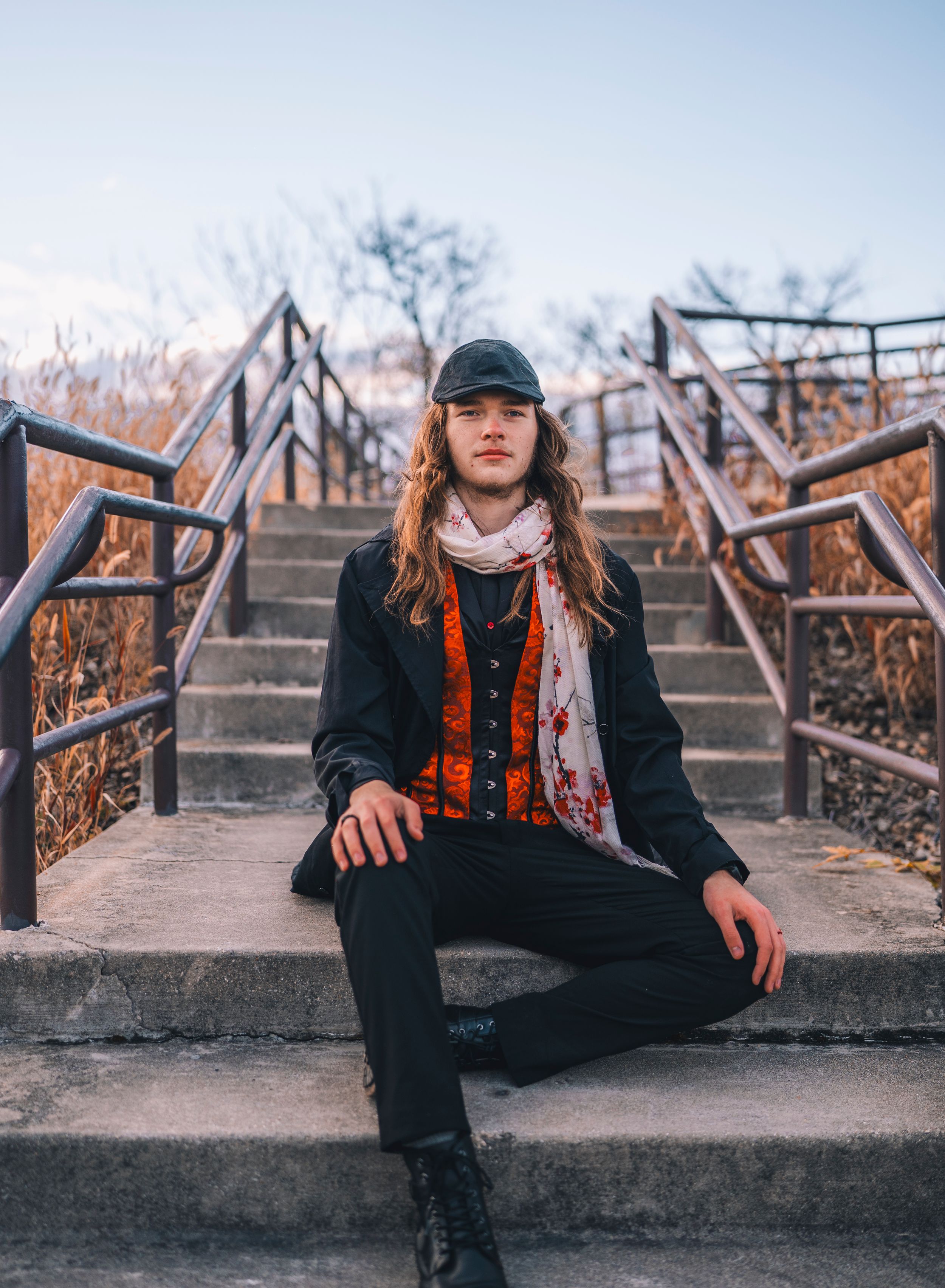 A person in black coat and orange vest sits on concrete steps in an outdoor urban setting during autumn.