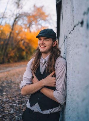 A portrait in autumn showing someone in casual attire with a cap leaning against a wall with orange foliage in background.
