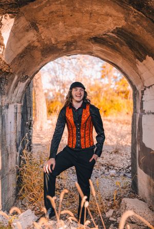Person in dark clothing and orange vest poses playfully under stone archways during golden autumn sunset.