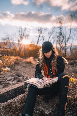 Person in dark clothing reads a large book while sitting on rocky ground during golden hour.